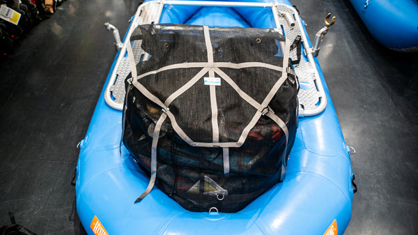 A black mesh cargo bag sitting in the back bay of a blue raft. Shown with the lid strapped closed and full of various rafting gear. 