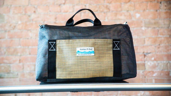 A black mesh boater's briefcase with d-rings, tan exterior pocket, and carry handles on a river table