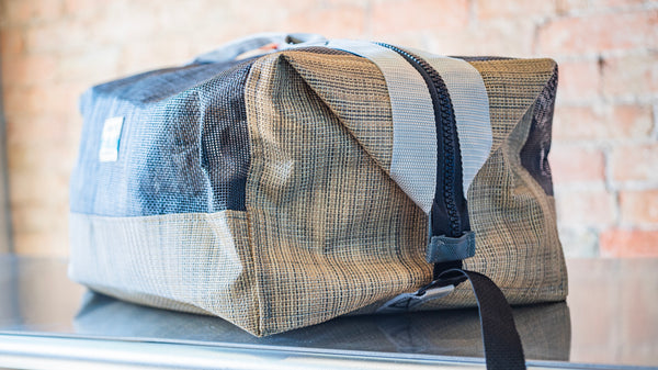 A tan and black mesh duffle bag showing the side closure clip and webbing tail. 