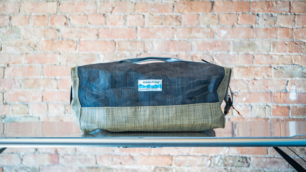A tan and black mesh duffle bag showing carry handles, side closures all on a river table. 