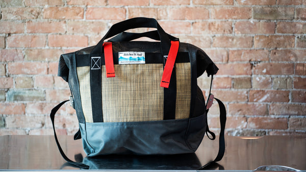 Tan and black roll top mesh bag showing a carry handle and cam straps on the side to secure the lid sitting on a river table. 