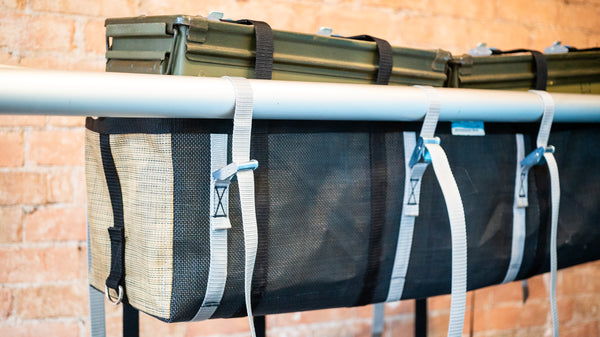A tan and black mesh bag with two rocket boxes intended for rafting strapped to the bag attached to a raft frame. 