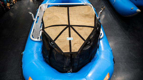 A tan and black mesh cargo bag sitting in the back bay of a blue raft. Shown with the lid strapped closed and full of various rafting gear. 