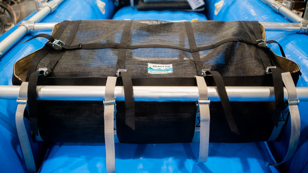 A front view of a tan and black mesh drop bag with a lid shown strapped to a raft frame on a blue raft. 