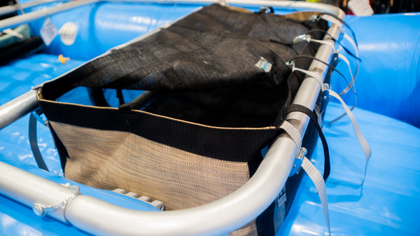 A tan and black mesh drop bag with a lid shown strapped to a raft frame on a blue raft. 