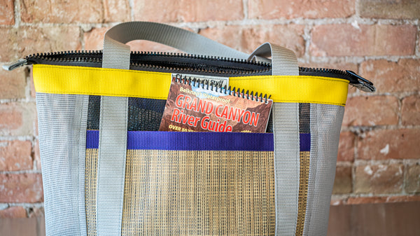 Tan and grey mesh zipper top tote with multi colored webbing on a river table with a river map in its exterior pocket.  