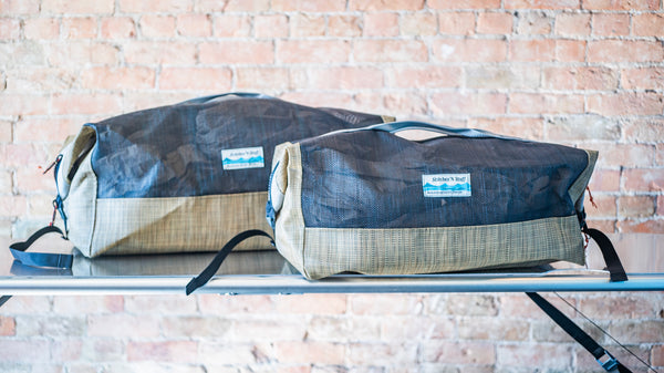 Size comparison between two tan and black mesh duffle bags showing carry handles, side closures all on a river table. 