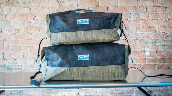 Size comparison between two tan and black mesh duffle bags showing carry handles, side closures all on a river table. 