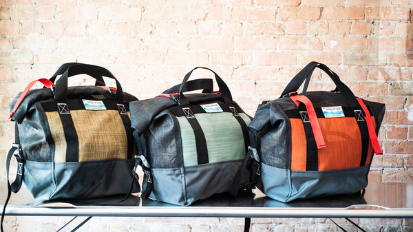 Three roll top mesh bags sitting on a river table showing the color options of tan, green, and orange.