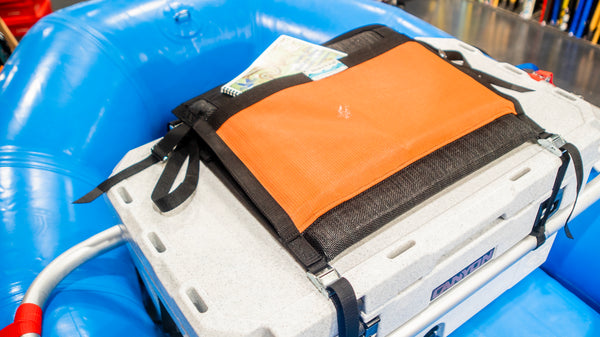 Top view of a padded orange and black mesh chair shown with the backrest down and displaying a river map in the built in pocket on a blue hyside raft. 