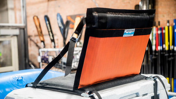 A padded orange and black chair strapped down to a rafting cooler with cam straps.
