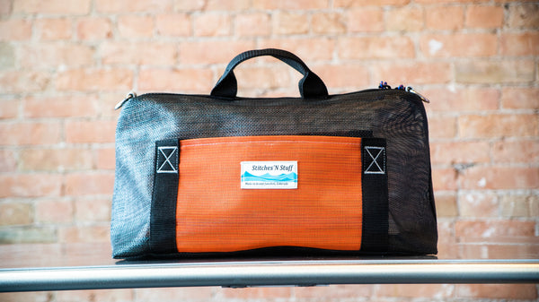 A black mesh boater's briefcase with d-rings, orange exterior pocket, and carry handles on a river table
