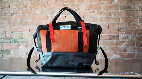 Orange and black roll top mesh bag showing a carry handle and cam straps on the side to secure the lid sitting on a river table. 