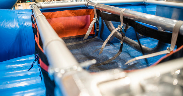 A Black and orange mesh drop bag with a zippered pocket in the front attached to a raft frame on a blue Hyside boat showing the direction of the cam buckle attachments.