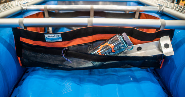 A Black and orange mesh drop bag with a zippered pocket in the front attached to a raft frame on a blue Hyside boat. Contents of the zippered pocket are a sand stake and a water bottle. 