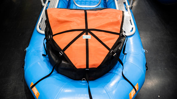 A orange and black mesh cargo bag sitting in the back bay of a blue raft. Shown with the lid strapped closed and full of various rafting gear. 