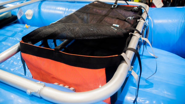 An orange and black mesh drop bag with a lid shown strapped to a raft frame on a blue raft. 