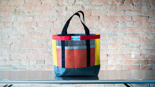 A multi colored mesh tote showing carry handles  and front exterior pocket shown on a river table. 