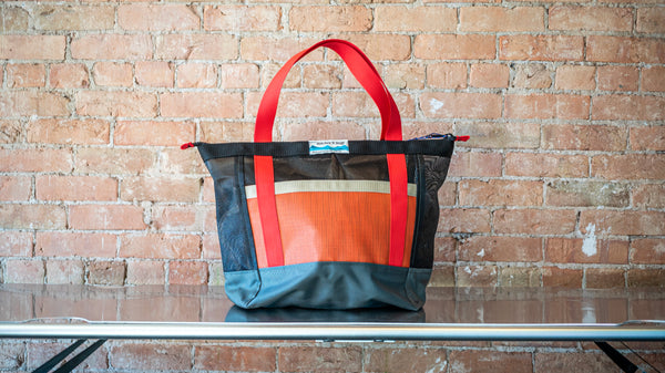Orange and black mesh zipper top tote with multi colored webbing on a river table. 