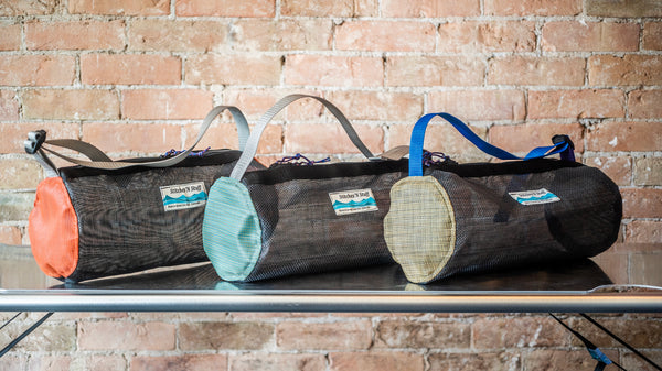 Three small mesh cylindrical duffle bags on a river table. Bags have a shoulder strap, one orange, one green, one tan. 