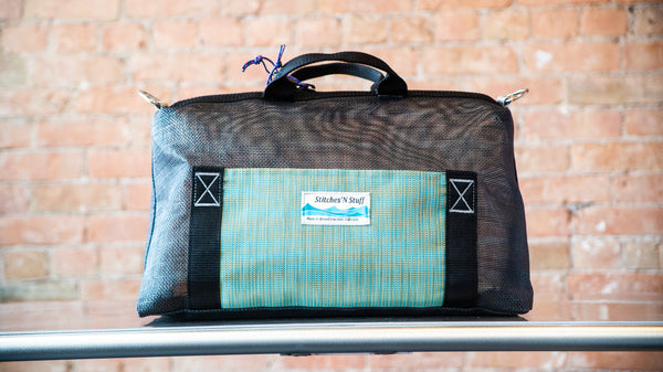 A black mesh boater's briefcase with d-rings, green exterior pocket, and carry handles on a river table