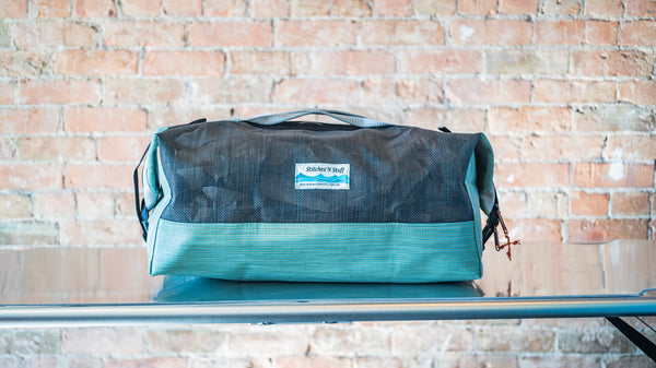 A green and black mesh duffle bag showing carry handles, side closures all on a river table. 