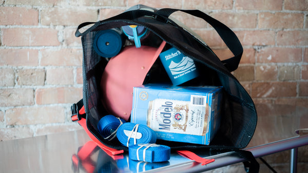Black mesh roll top bag showing the lid open and filled with water bottles, whitewater helmets, cam straps, and drinks. 