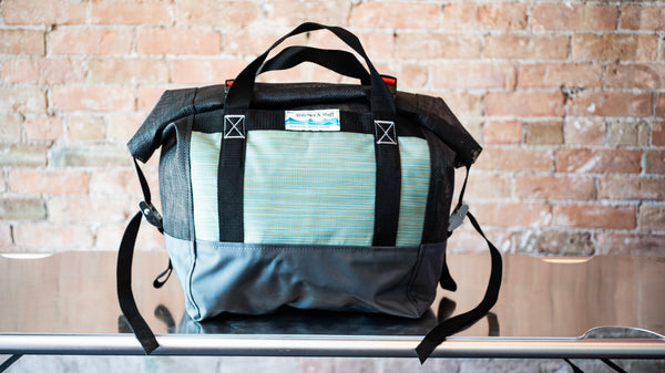 Green and black roll top mesh bag showing a carry handle and cam straps on the side to secure the lid sitting on a river table. 