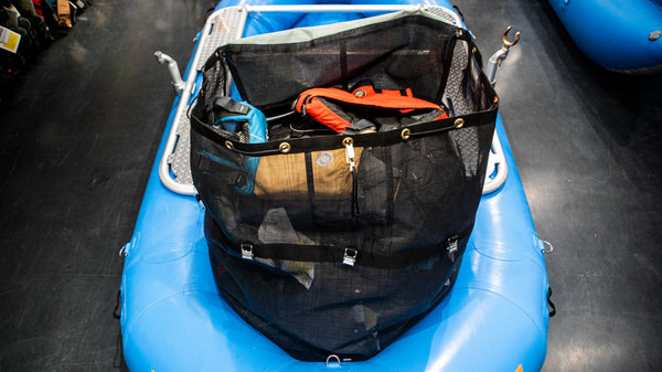 A green and black mesh cargo bag sitting in the back bay of a blue raft. Shown with the open and full of various rafting gear. 