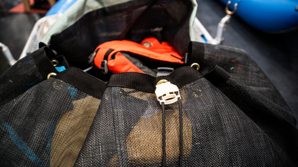 A green and black mesh cargo bag sitting in the back bay of a blue raft. Shown with the lid cinched closed and full of various rafting gear. 