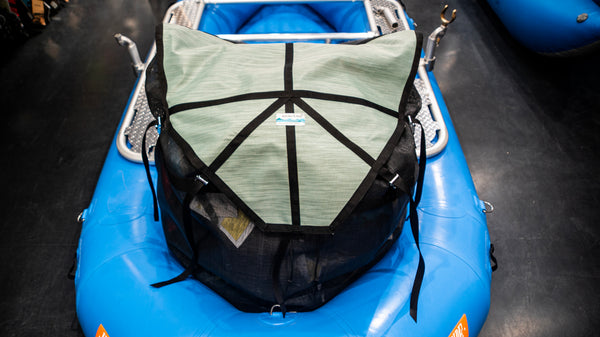 A green and black mesh cargo bag sitting in the back bay of a blue raft. Shown with the lid strapped closed and full of various rafting gear. 