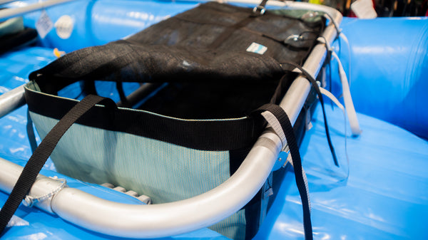 A green and black mesh drop bag with a lid shown strapped to a raft frame on a blue raft. 