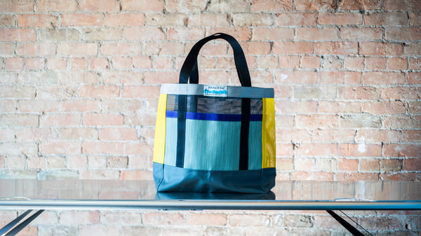 A multi colored mesh tote showing carry handles  and front exterior pocket shown on a river table. 