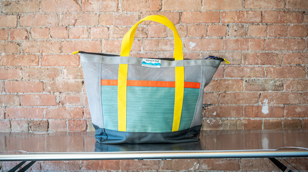 Green and grey mesh zipper top tote with multi colored webbing on a river table. 