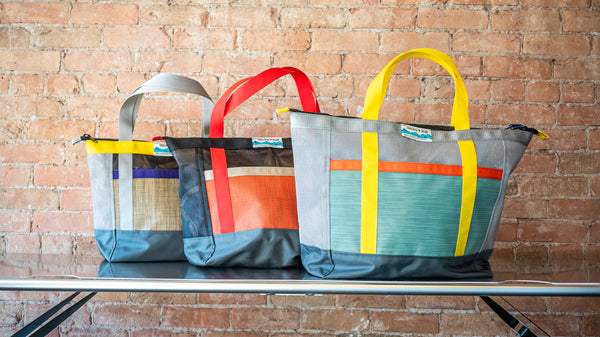 Three tote bags, one green, one red, one tan with different colored webbing on a river table.