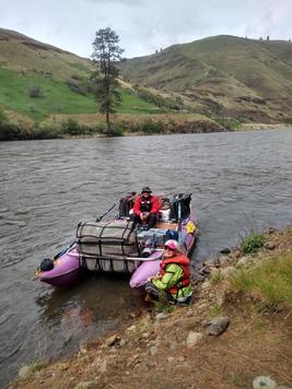Purple cataraft on a river bank with rower on rowers seat and mesh bag in the front of the frame. Hills and trees occupy the river bank. 